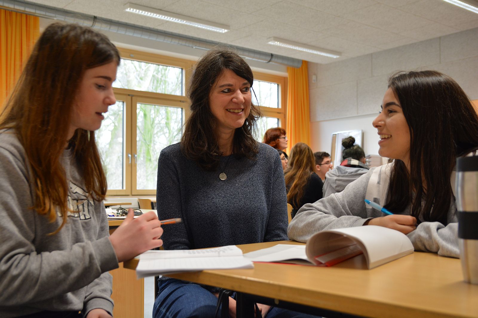 Betty Kolodzy sitzt grinsend zwischen zwei Schülerinnen, Texte vor ihnen liegend