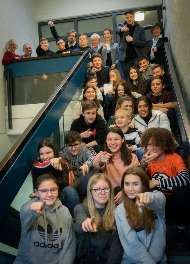 Gruppenfoto von Schüler*innen der Oberschule in den Sandwehen auf einer Treppe, alle zeigen mit dem Finger in die Kamera, oben stehen Lehrer*innen und Heike Müller